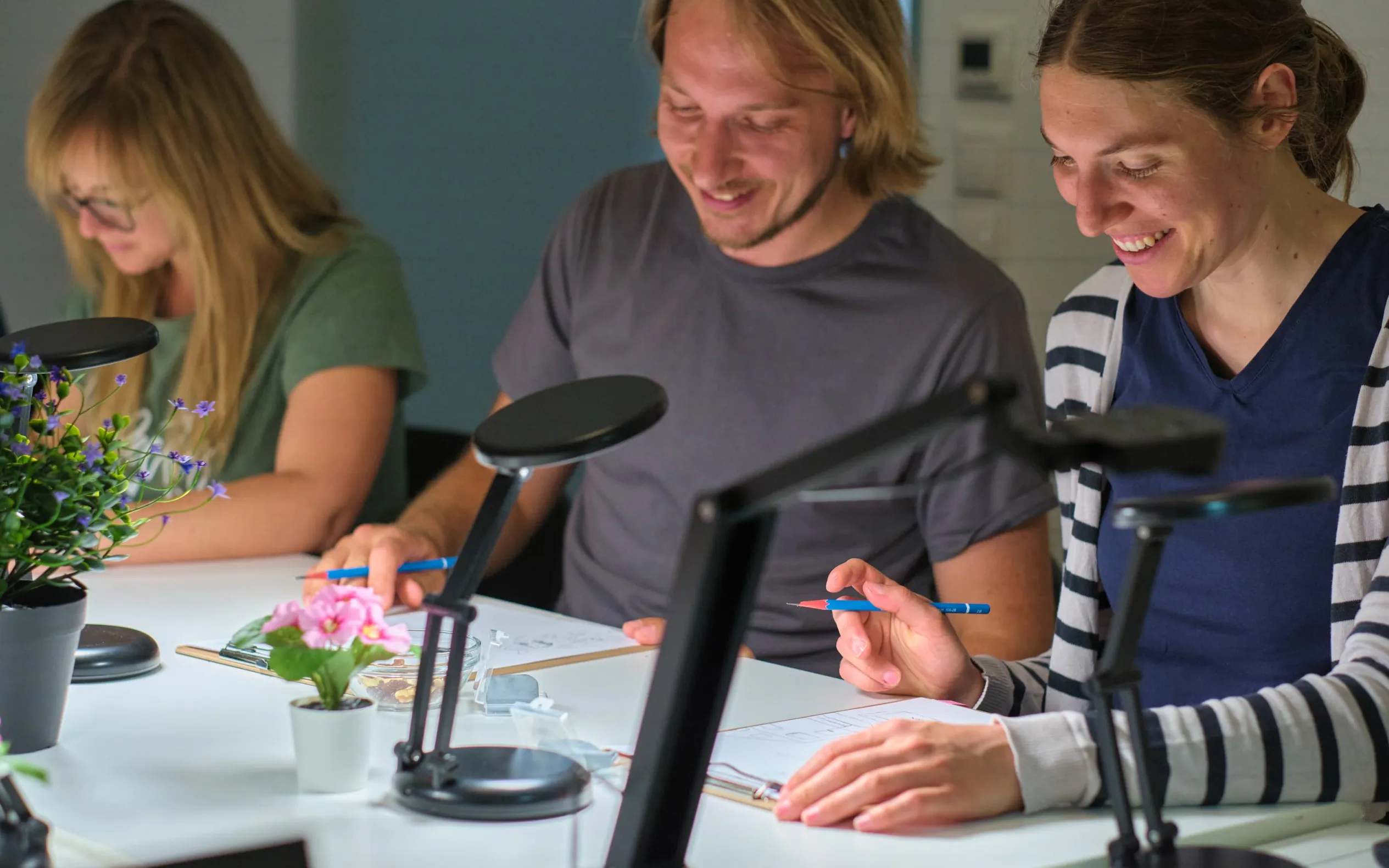 Drei Kursteilnehmer:innen zeichnen konzentriert unter Tischlampen